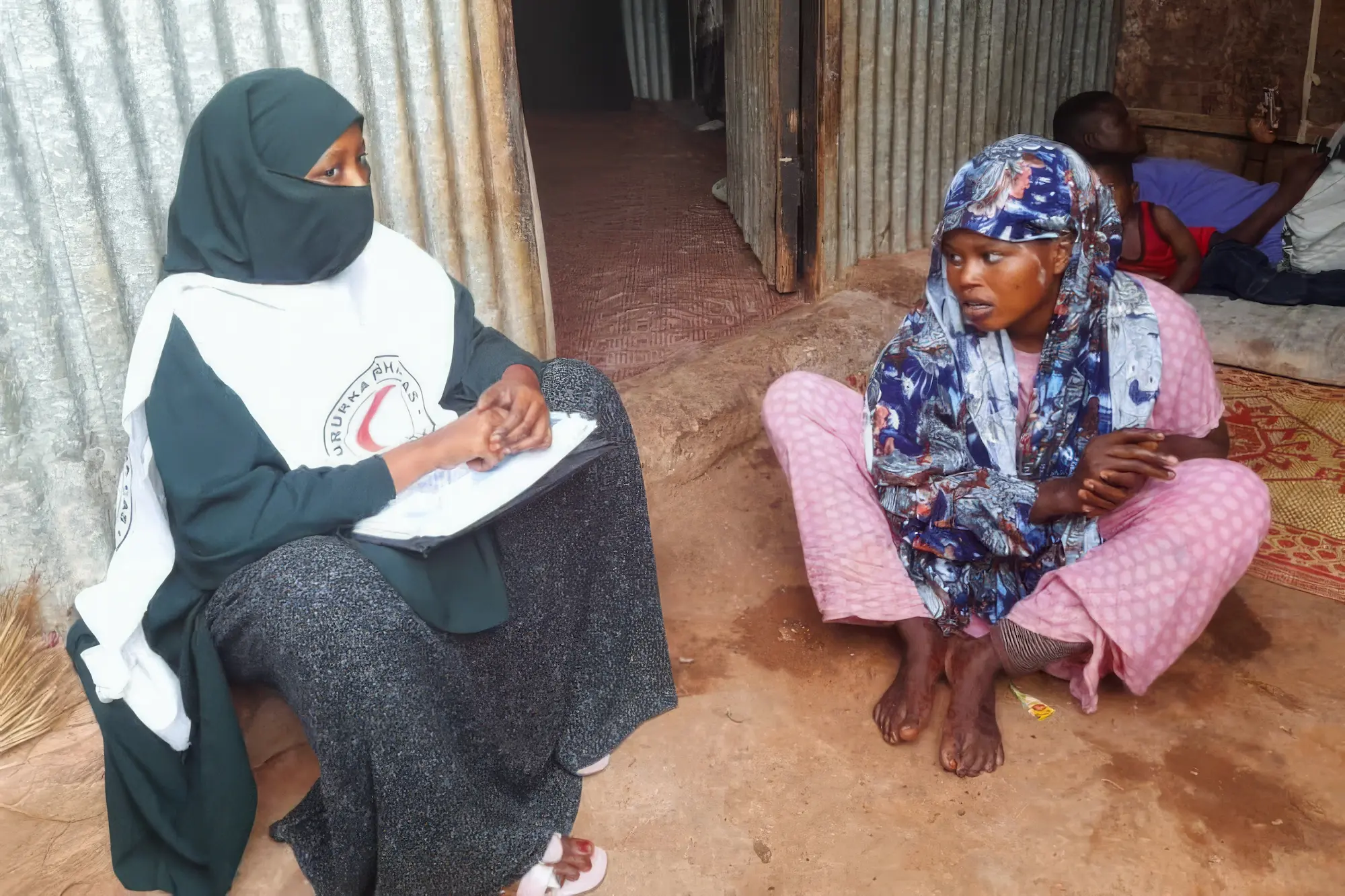 An IFRC staff member talking to a lady outside their house, sitting on the ground