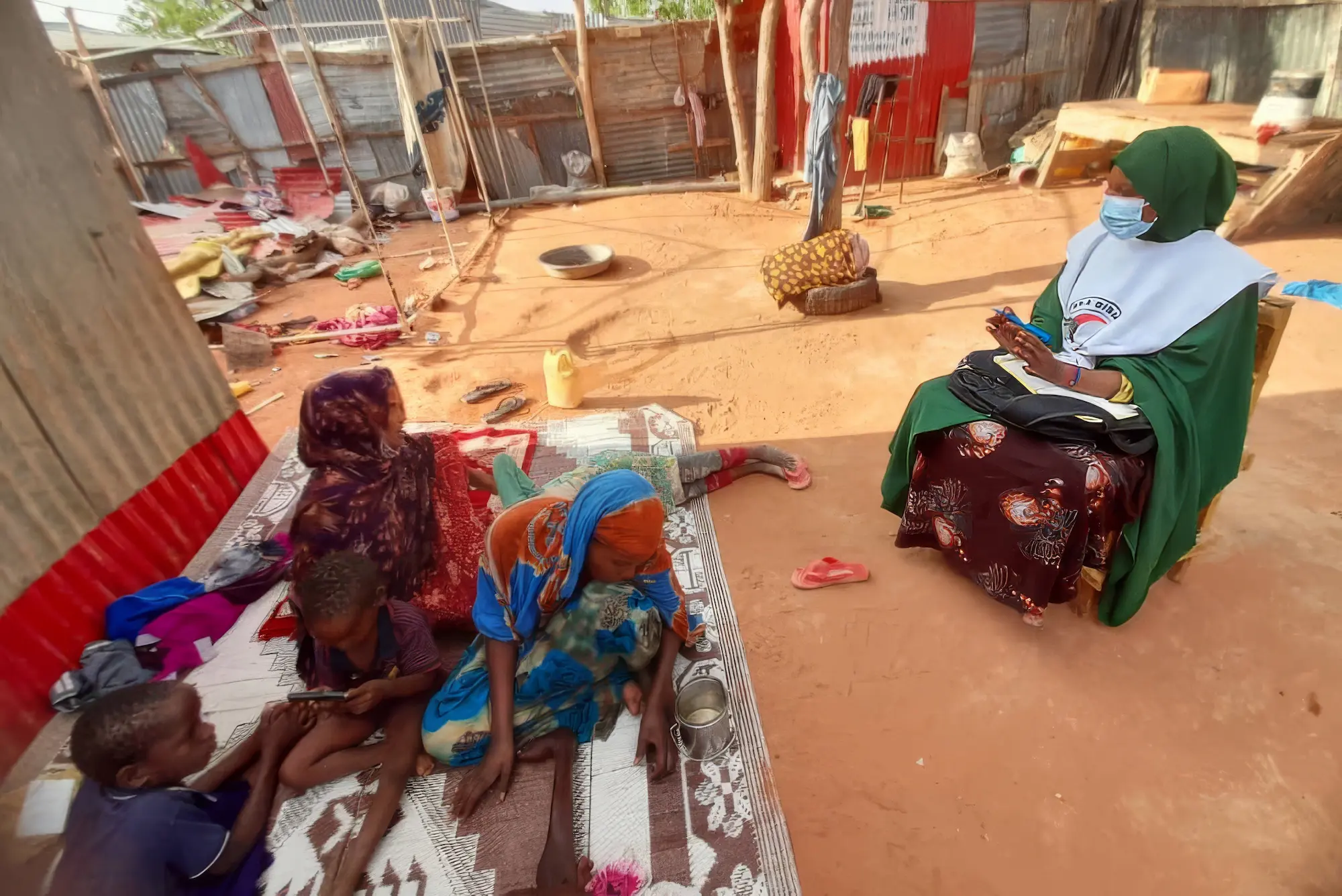 IFRC staff member sits on a chair, wearing a mask, talking to a mother and her two children outside their house, sitting on a rug