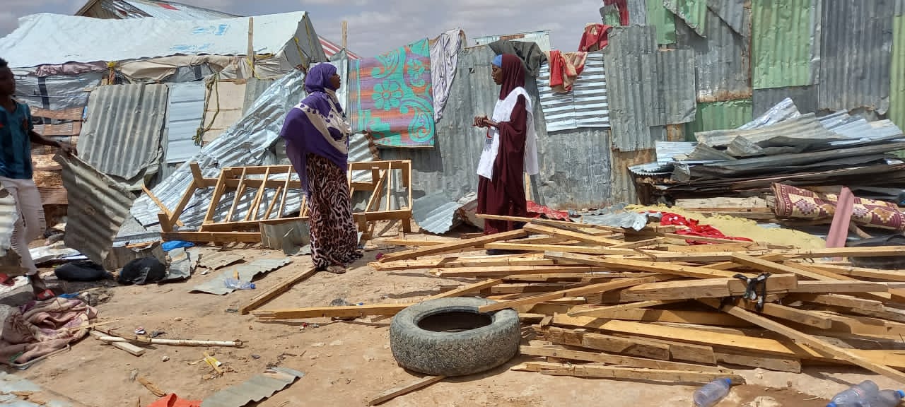 Two women talking amongst debris of wooden planks, corrugated metal and an old tyre