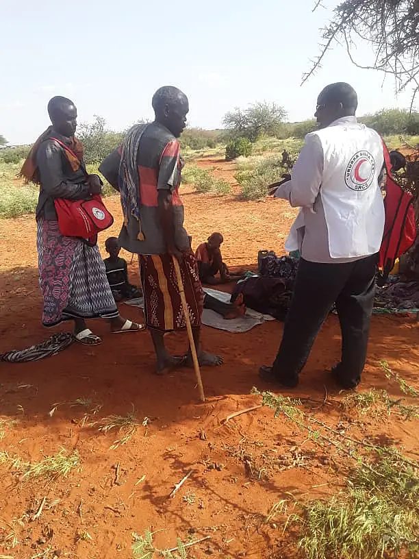 An IFRC staff member talking to two villagers under the shade of a tree. One is leaning on his staff