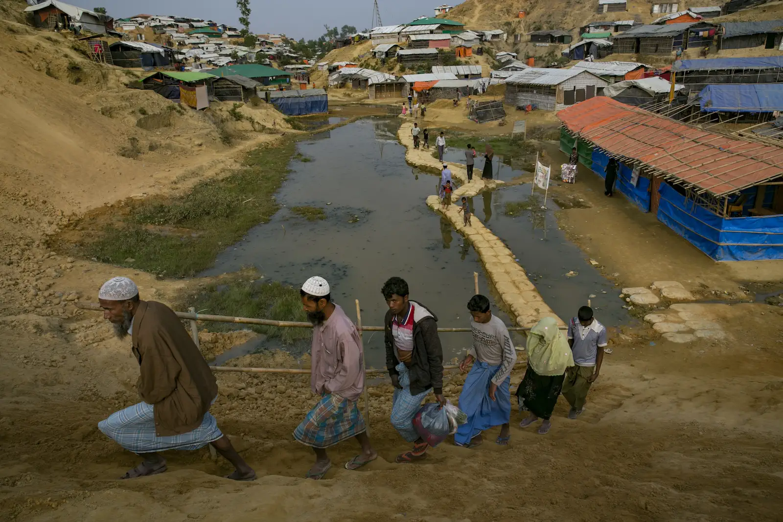 Refugees walking over a path through a pool next to the camp. Five males, and one female are in the foreground climbing a hill