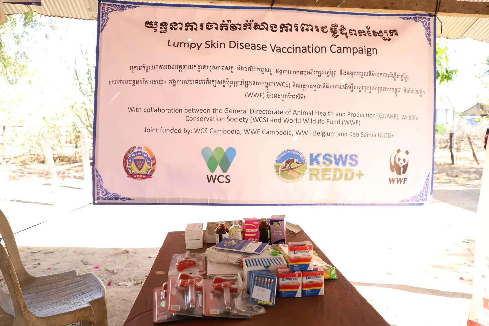 A vaccination campaign poster hanging above a table of medical supplies, bright sunshine behind