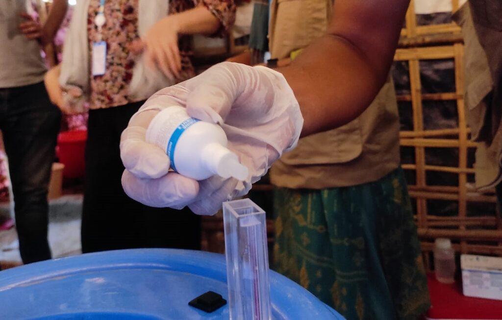 A latex gloved hand squeezing a pipette of chemicals into a sample of water