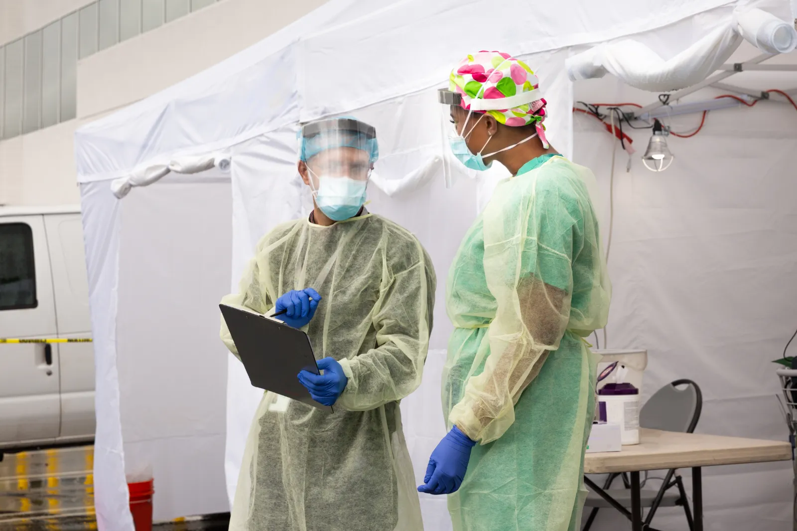 Two heathcare workers in full PPE discussing information on a clipboard, outside a tent
